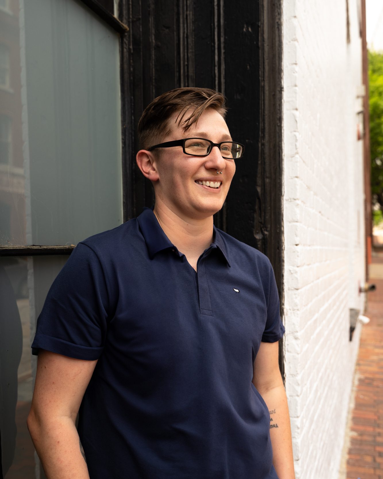 A white nonbinary person with short brown hair and black glasses smiles and looks to the side of the camera